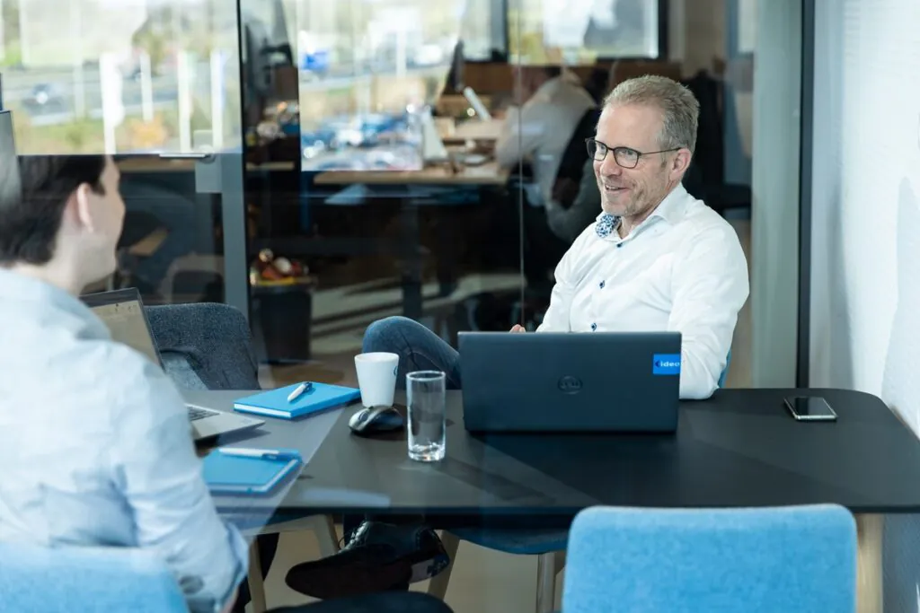 Two professionals discuss field service management with a DELL laptop (ideo sticker) between them in a modern office setting.
