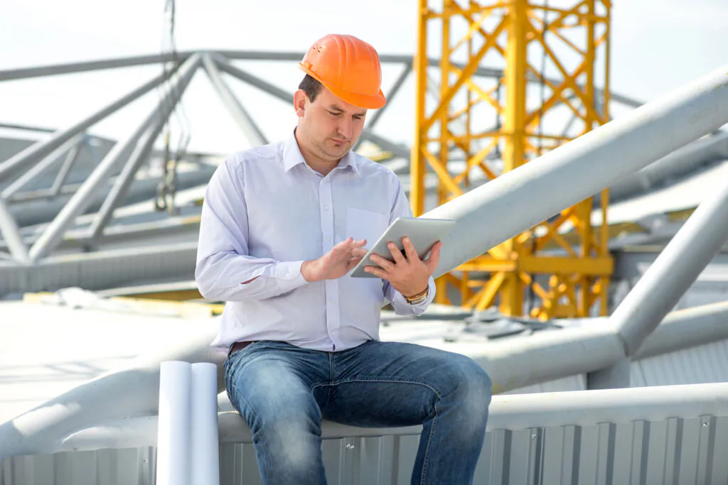 Man in orange hard hat sitting, interacting with a tablet on a construction site. Metal beams and a yellow crane are visible in the background, suggesting field service management.