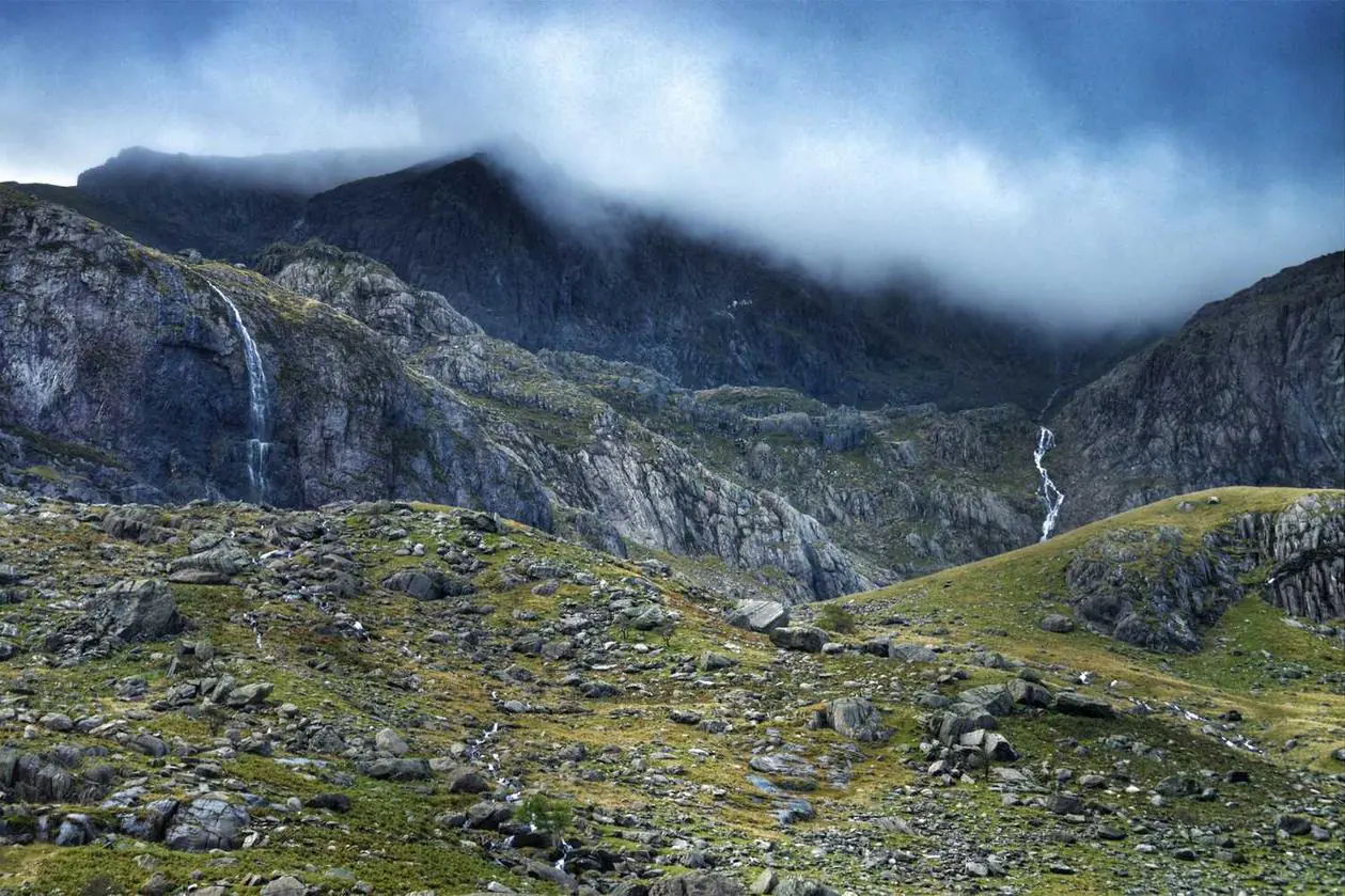 Rugged mountain landscape featuring cascading waterfalls and rocky slopes, valuable natural **assets** under a cloudy, misty sky.
