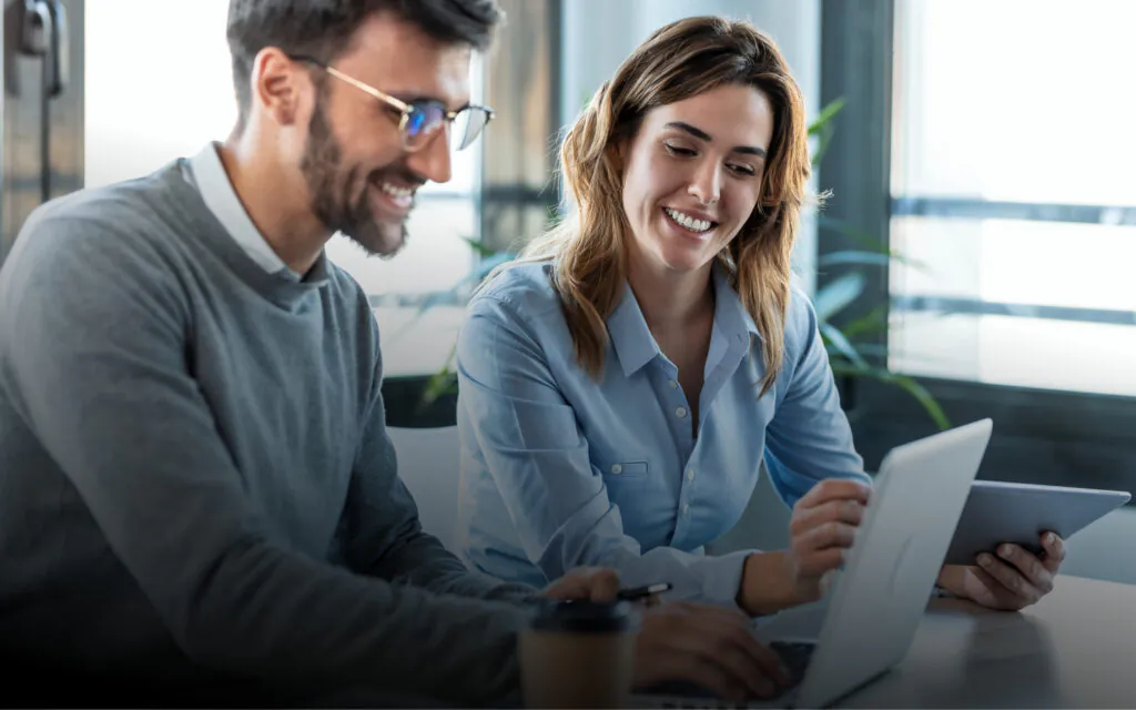 Two smiling professionals use sap technology, collaborating on a laptop and tablet at a modern office desk.