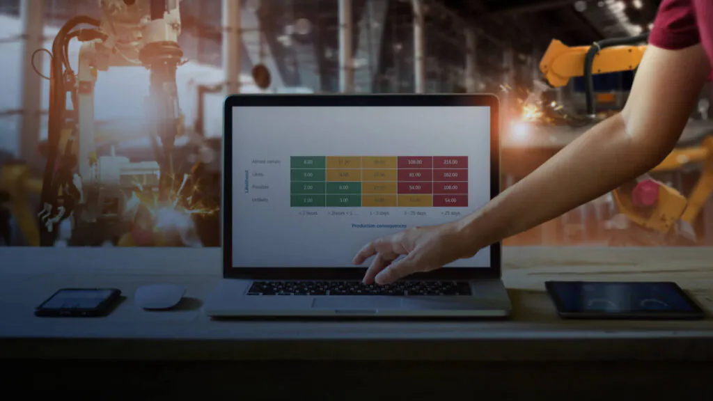 A hand points at a laptop displaying a risk matrix for asset management, with industrial robotic arms welding in the blurred background. **Text:** Likelihood Almost certain Likely Possible Unlikely Production consequences < 2 hours > 2hours < 1... 1 - 3 days 3 - 25 days > 25 days 4.00, 12.00, 36.00, 108.00, 216.00 3.00, 9.00, 27.00, 81.00, 162.00 2.00, 6.00, 18.00, 54.00, 108.00 1.00, 3.00, 9.00, 27.00, 54.00