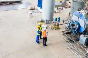 Three engineers in hard hats and safety vests inspect large industrial equipment, including a blue boiler, in a vast factory. They are engaged in **asset management**.