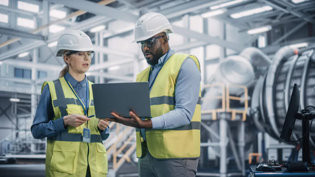 Two engineers in hard hats and vests review data on a laptop, discussing **asset management** in a modern industrial facility. (Text on helmets: AERO)