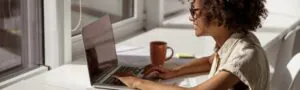 A smiling woman with curly hair and glasses types on a laptop at a sunny white desk by a window, with a mug and papers nearby.