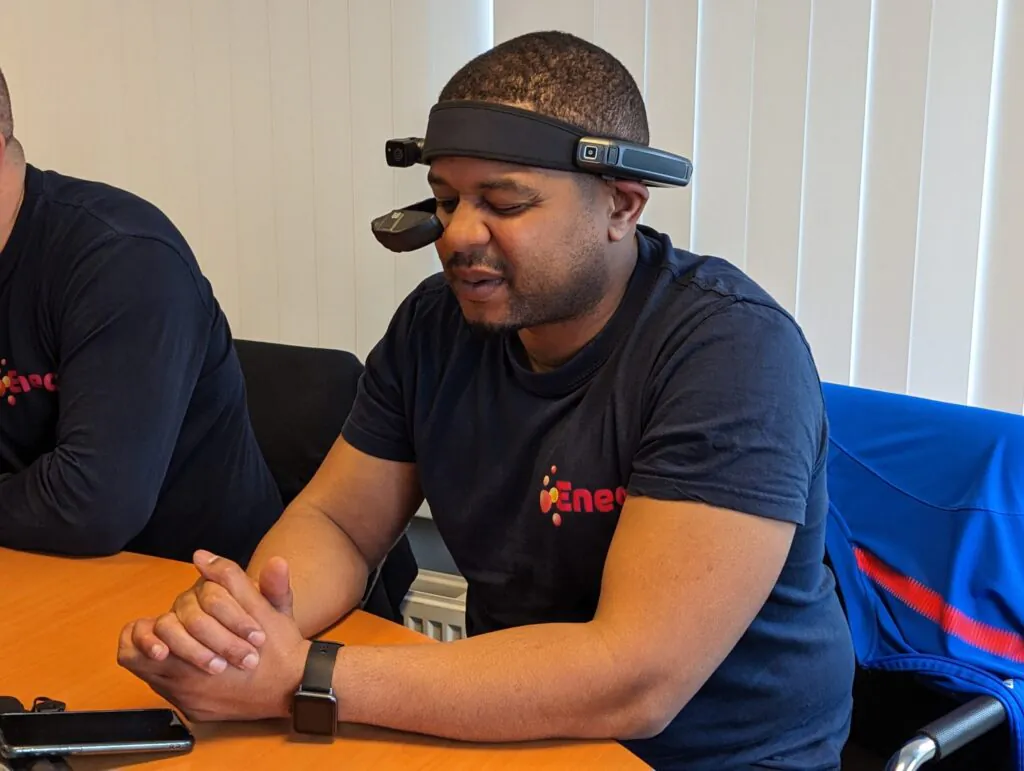 A man wearing a head-mounted device and dark blue 'Eneco' t-shirt sits with clasped hands at a wooden table, for potential use in **field service management**.