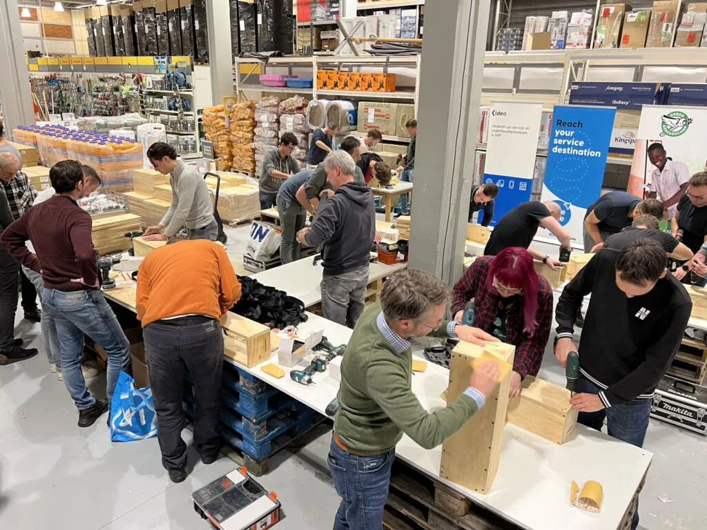 Multiple individuals assemble wooden boxes using drills at workstations within a brightly lit warehouse store. This workshop could enhance **field service management** skills. **Transcribed text:** IdeO, Verbeter uw service- en onderhoudsprocessen in SAP, Reach your service destination, Kingspan, GERED GEREEDSCHAP, Voor de Klus, PRIJS GARANT!, Makita.