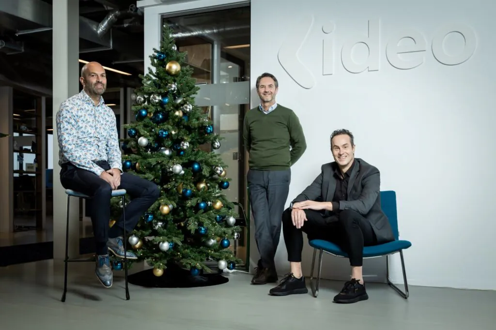 Three smiling men pose in an office. One sits on a stool, one stands by a decorated Christmas tree, and one sits in a chair. Text: Kideo.