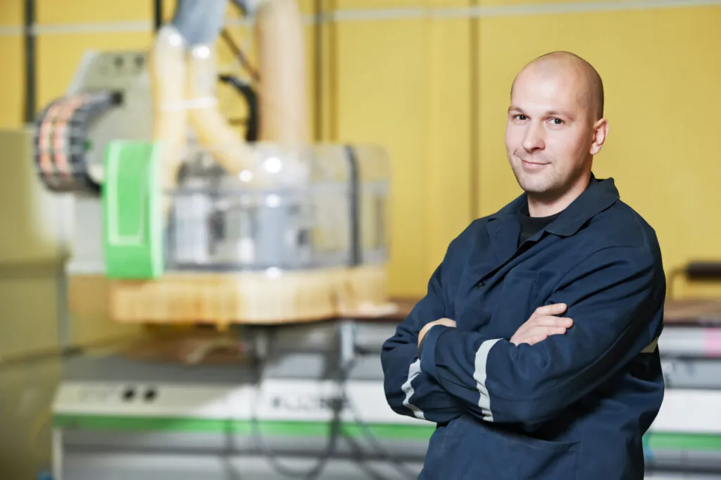 A bald man in a dark blue work jacket, likely involved in field service management, smiles with crossed arms in a factory setting with blurred industrial machinery.