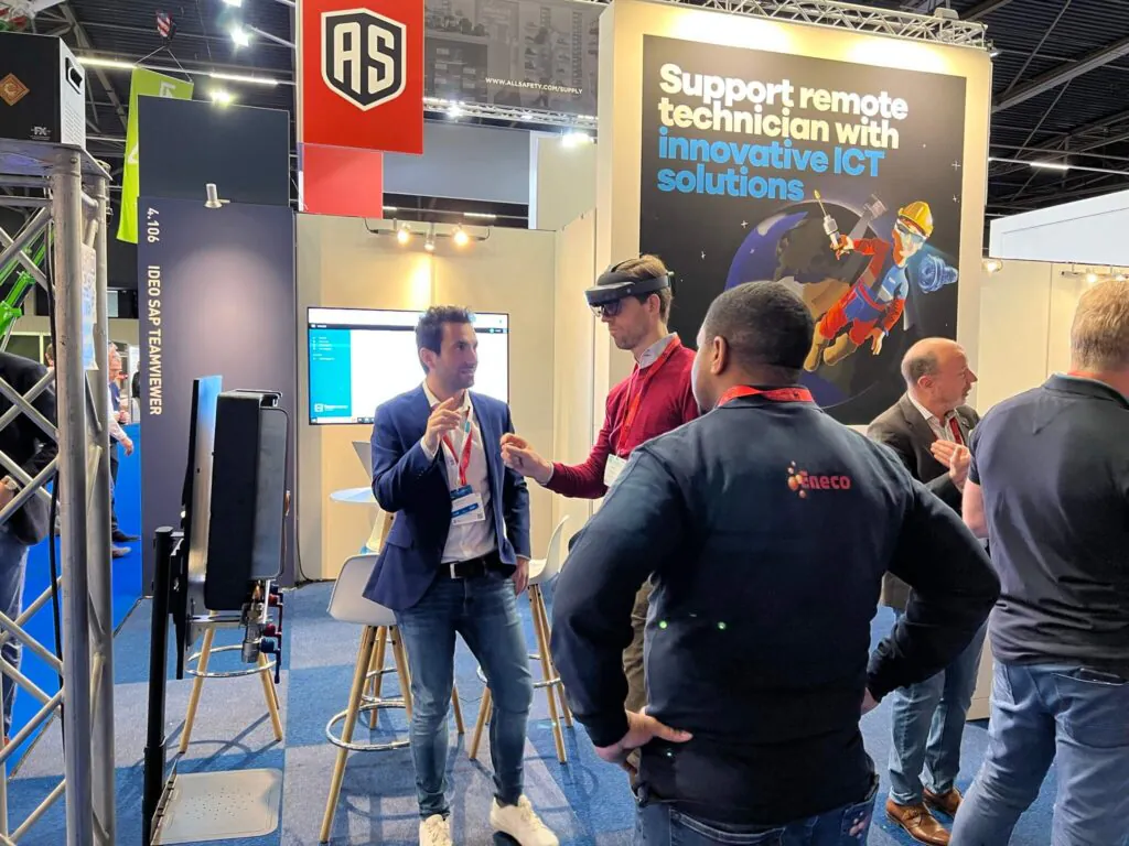 Men discuss a demonstration of augmented reality for **field service management** at a tech exhibition booth. Text: "4.106 IDEO SAP TEAMVIEWER", "Support remote technician with innovative ICT solutions", "AS", "WWW.ALLSAFETY.COM/SUPPLY", "Eneco".