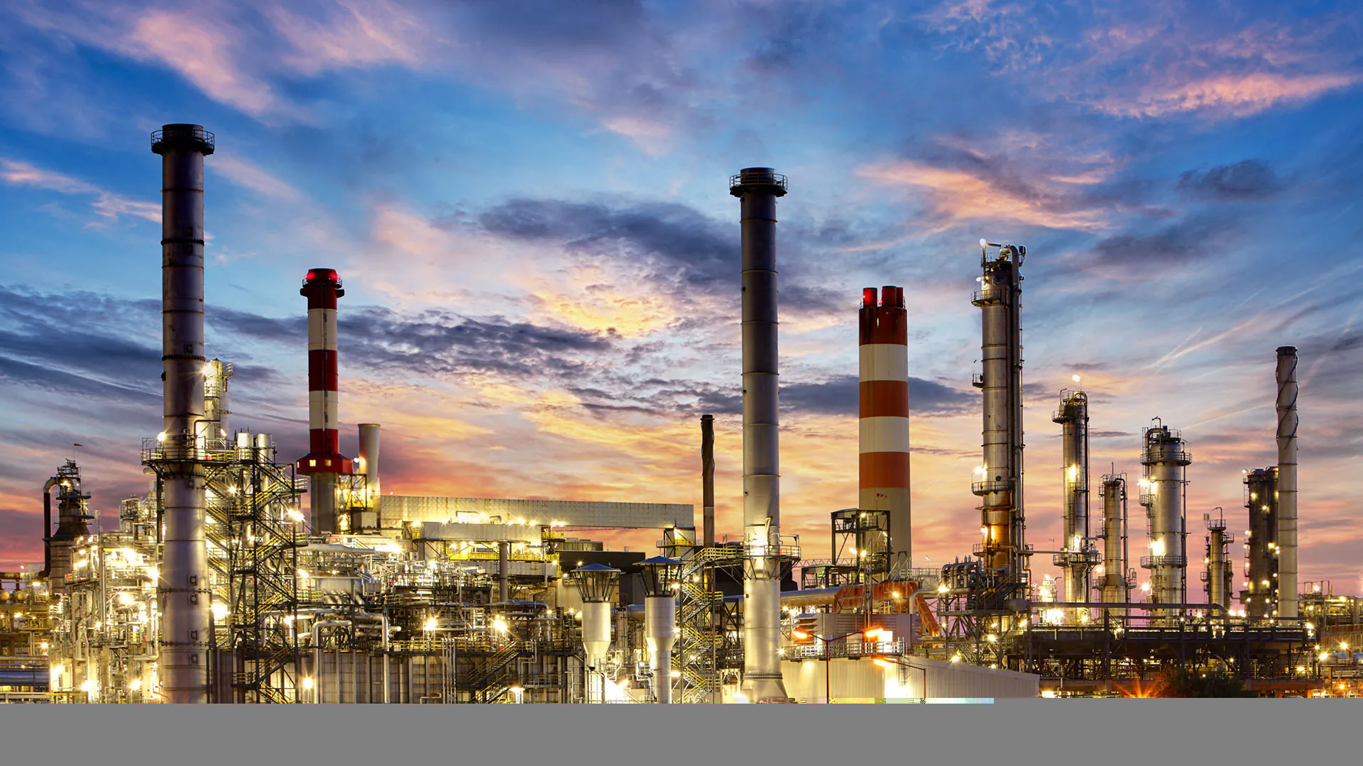 An expansive industrial refinery, with numerous illuminated towers and pipes, operates under a dramatic blue and orange twilight sky, showcasing complex asset management.