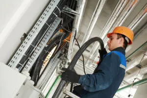 A technician in an orange hard hat routes a thick black cable through metal trays filled with wires, managing installations in an industrial ceiling. This demonstrates **field service management**.