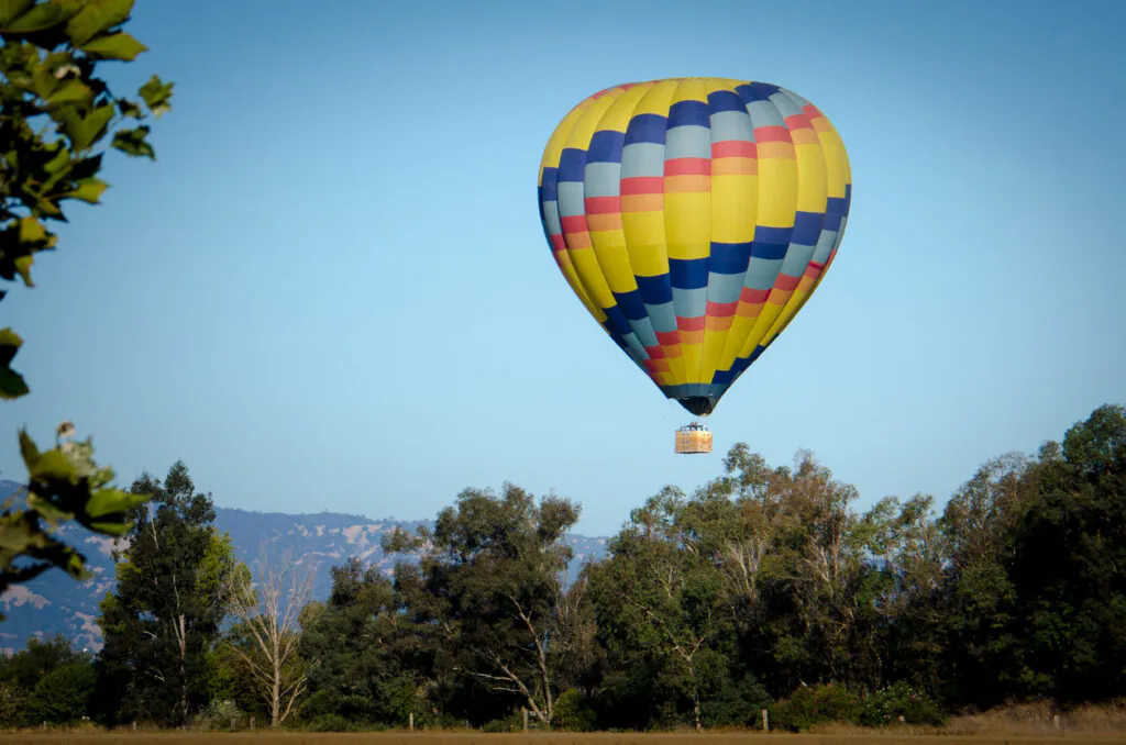 A multicolored hot air balloon floats above a treeline with distant hills under a clear blue sky.