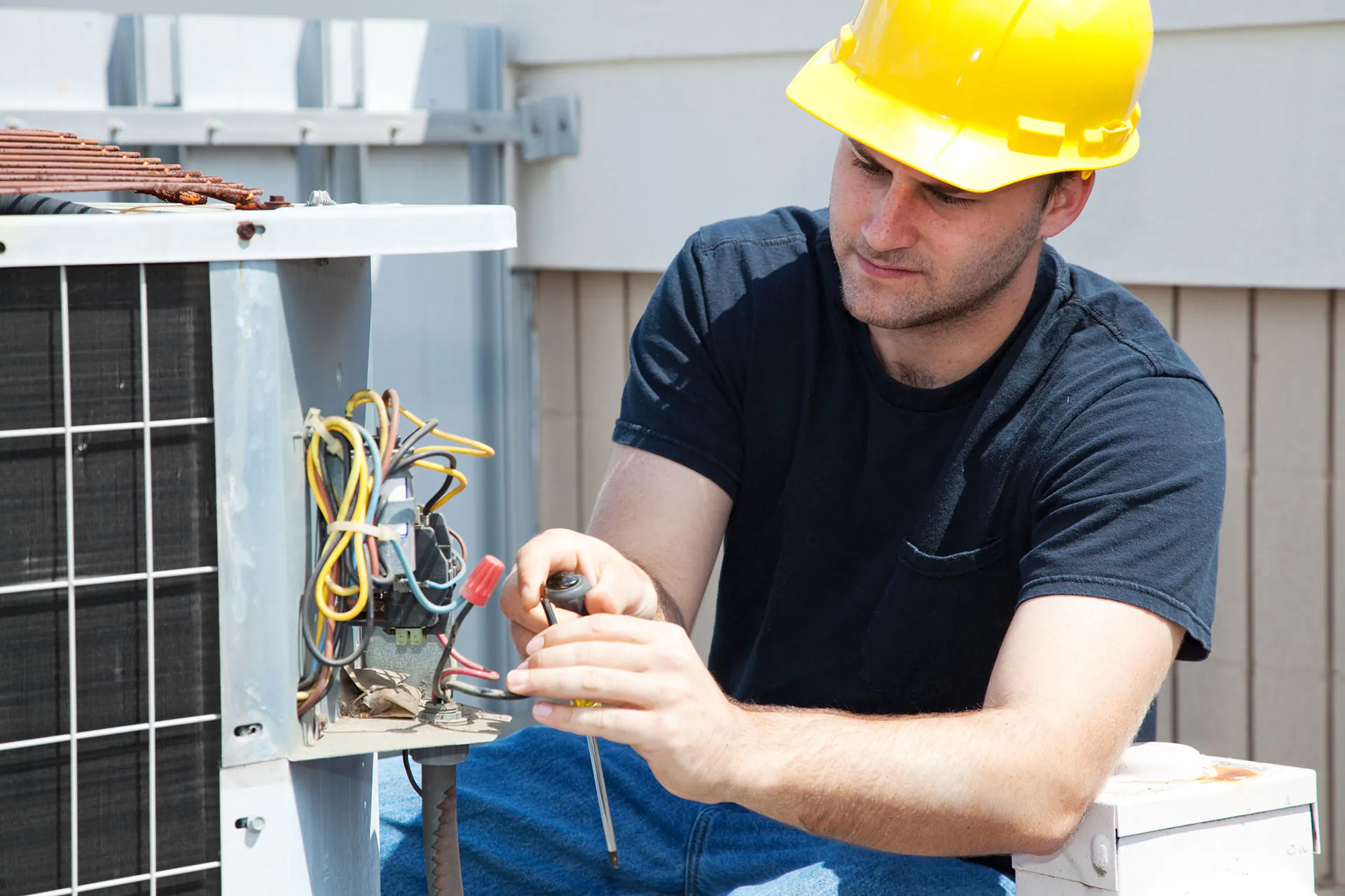 Male technician in a yellow hard hat intently repairs an outdoor air conditioning unit, adjusting its complex electrical wiring with a screwdriver for field service management.