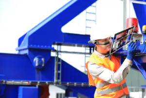 A field service management technician wearing smart glasses and a safety vest uses a tablet (AVION, 00:00:23) for remote assistance on large blue industrial machinery.