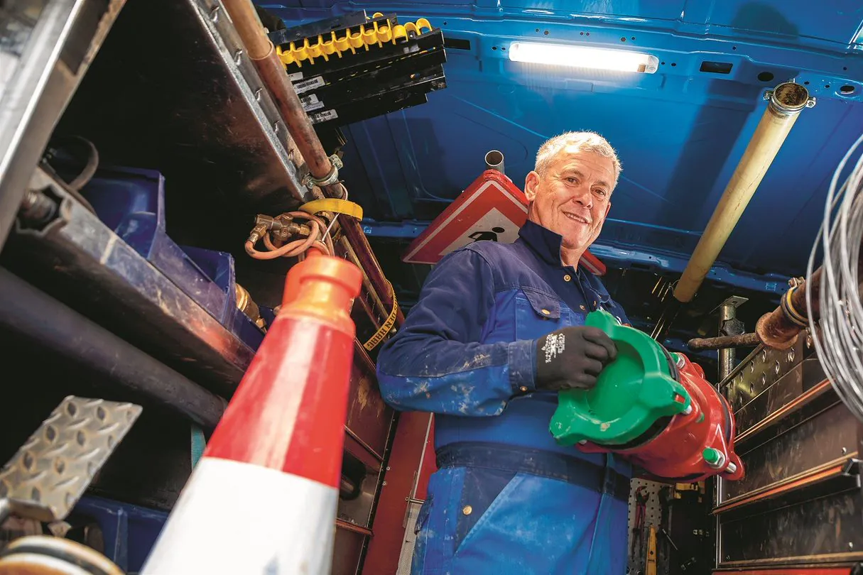 Smiling field service worker holds a red and green tool, looking at the camera, within a tool-laden van. Text: SCHILLER WEG, DEN HHBC.