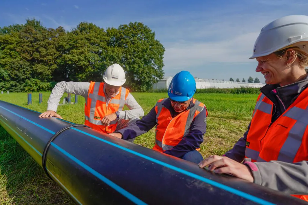 Three field service management workers (Van Gelder & Van Gelder, Vitens, min Hanab) inspect a black pipe with blue stripes (SDR 11 110x10.0mm PE100 RC ISO 4427/EN 12201 2022) in a grassy field with trees and greenhouses.