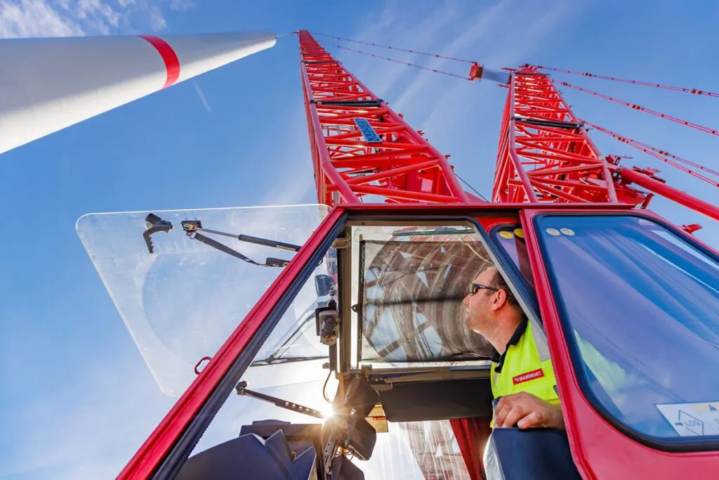 A crane operator in a 'MAMMOET' vest operates a red 'LIEBHERR' crane, looking up at a wind turbine tower being installed under a clear sky, an example of field service management.