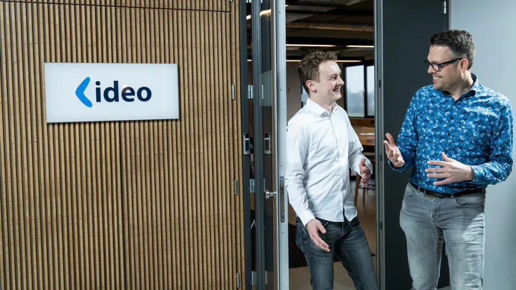 Two men discuss SAP technology, smiling and conversing in a modern office doorway. A backlit sign reads "ideo" on a textured wall.