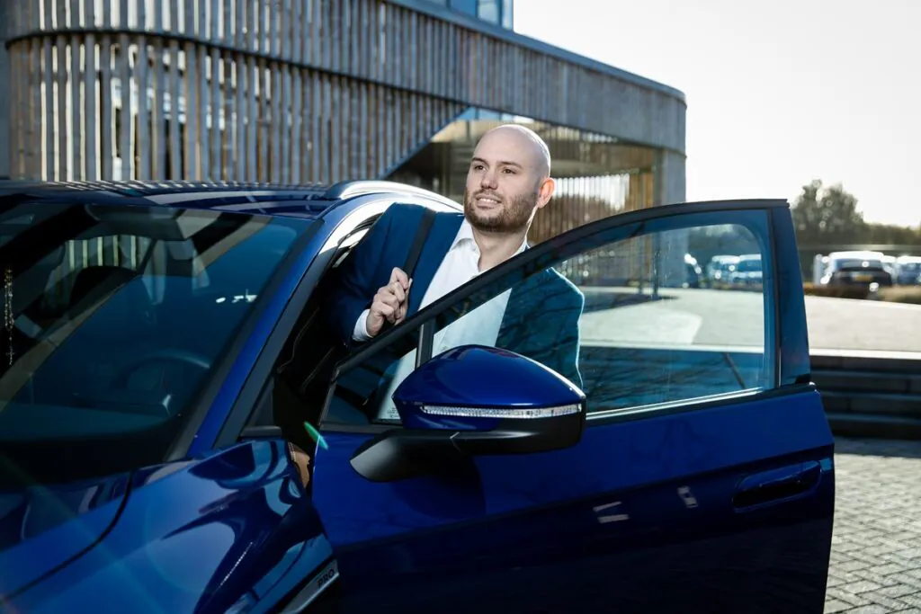 A bald, bearded man in a suit, likely for field service management, steps out of a blue "PRO" car, looking right, in a sunny parking lot by a modern building.