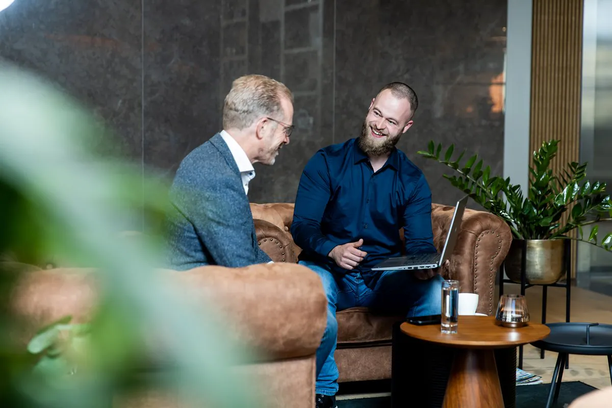 Two men discuss SAP FSM solutions, smiling on a brown leather sofa. One holds a laptop in a modern office lounge with plants.