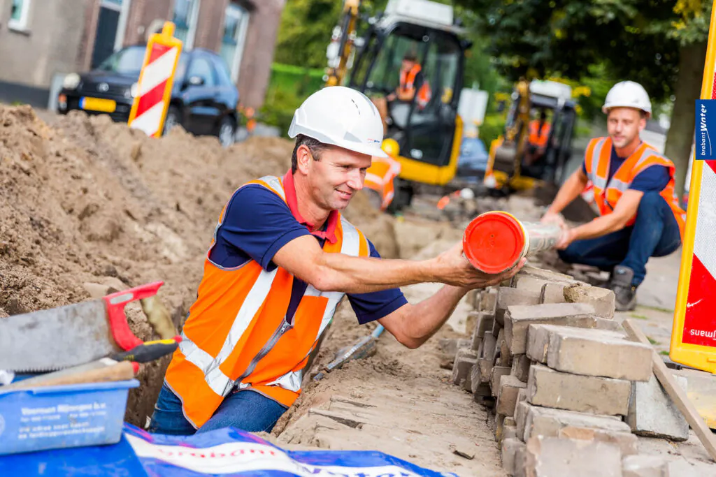 A **field service management** worker, wearing a "brabant Water" hard hat, places a "MÜLLER PE100 SDR11 NET" pipe into a trench. A colleague and excavator work at the construction site, with signs ("brabant Water", "suw...") and a container ("Voerman Nijmegen b.v. [...]"). A sheet on the ground reads "brabant Water water".