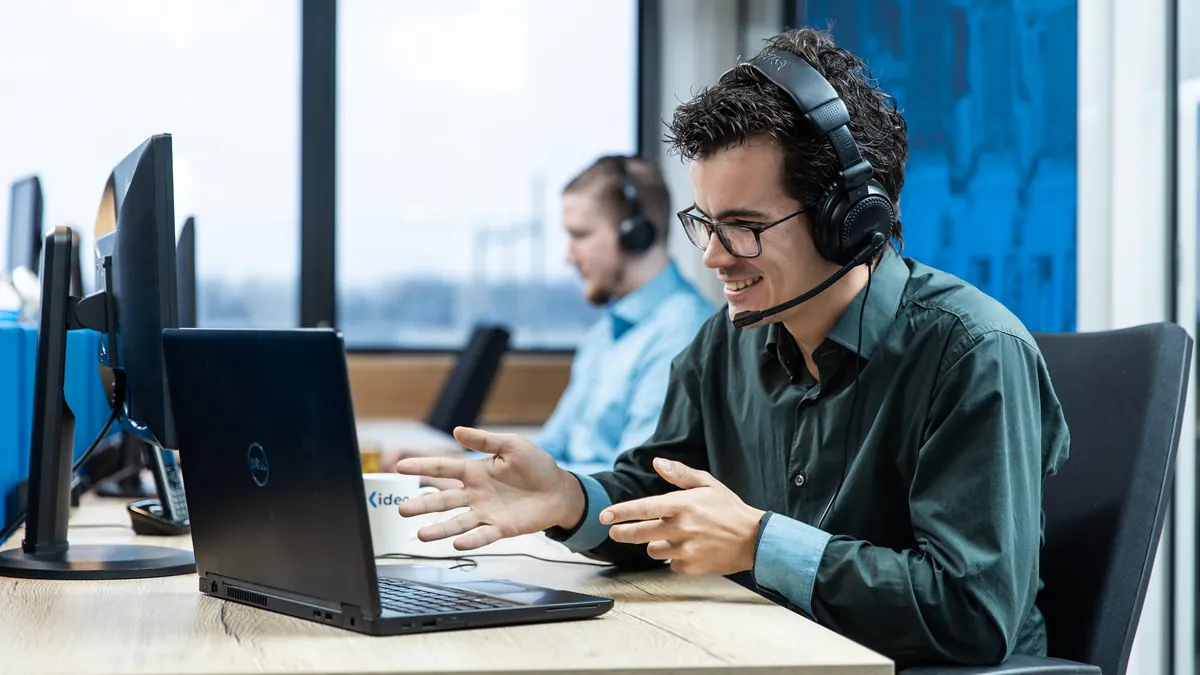 A young man with Jabra headset and glasses smiles, gesturing at a DELL laptop screen at an office desk. A mug reads "idea". He offers SAP FSM support.