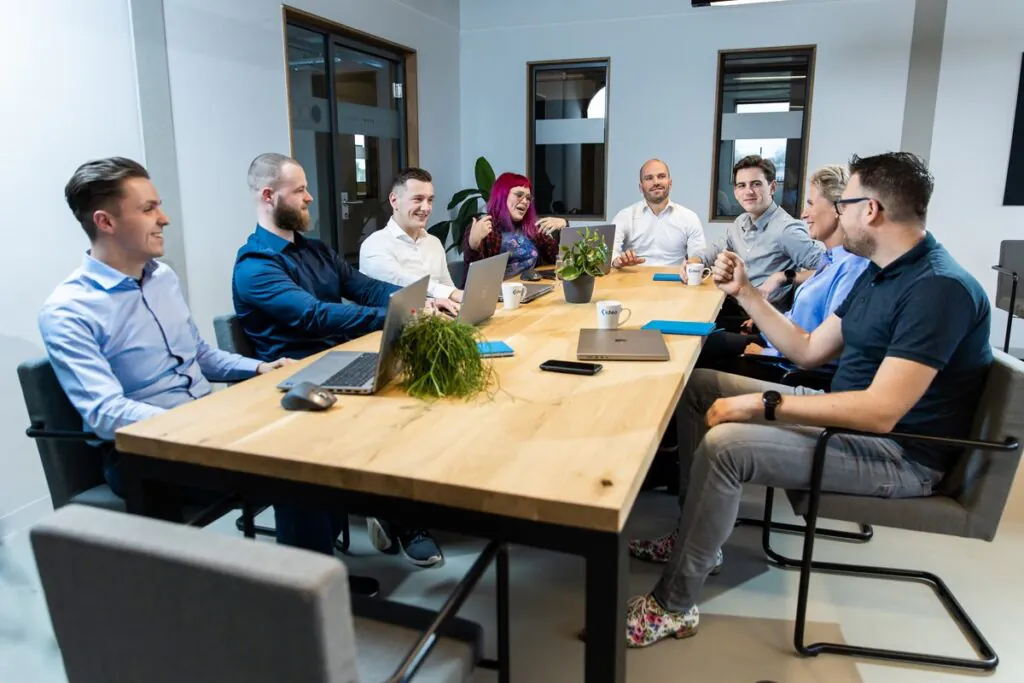 Eight diverse professionals are smiling and discussing around a large wooden table with laptops in a modern office. Mugs read "‹ideo". Glass partition text: "GOOD PEOPLE", "GOOD PROJECTS", "OODO". This shows a team using **SAP technology**.