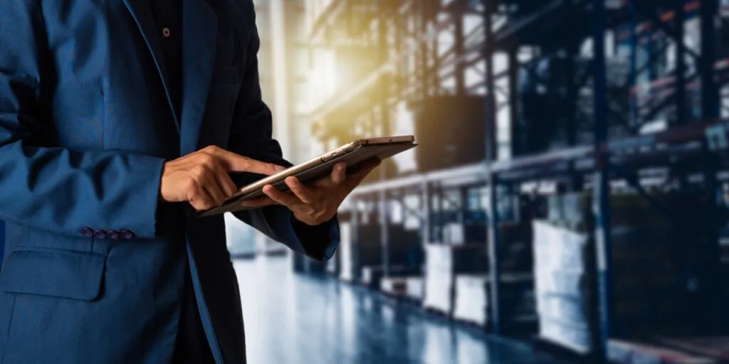 A person in a blue suit jacket uses a tablet for asset management, standing in a blurred, industrial warehouse with tall shelving.