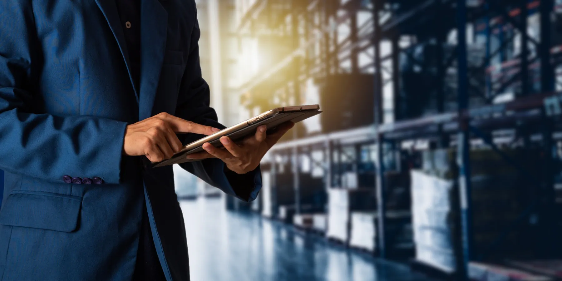 A person in a blue suit jacket uses a tablet for asset management, standing in a blurred, industrial warehouse with tall shelving.