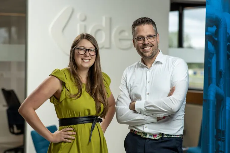 Smiling man and woman pose in an office. She wears a green dress with hands on hips; he, a white shirt with arms crossed. "idea" is embossed on the wall.