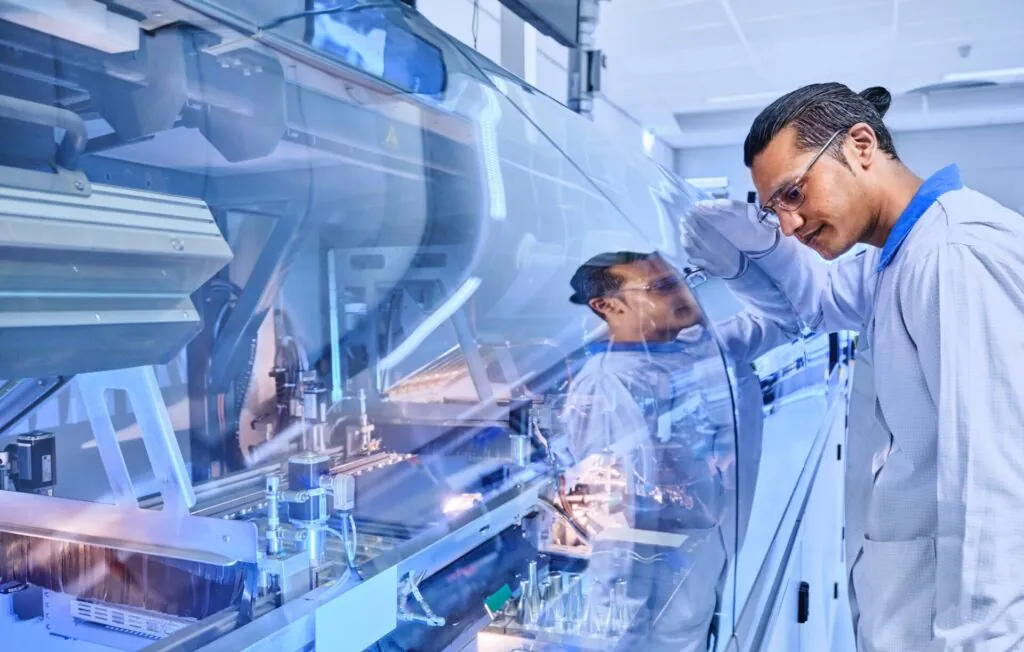 A technician in a lab coat and gloves inspects complex industrial machinery through a clear cover displaying a warning triangle. He is performing asset management in a clean room.