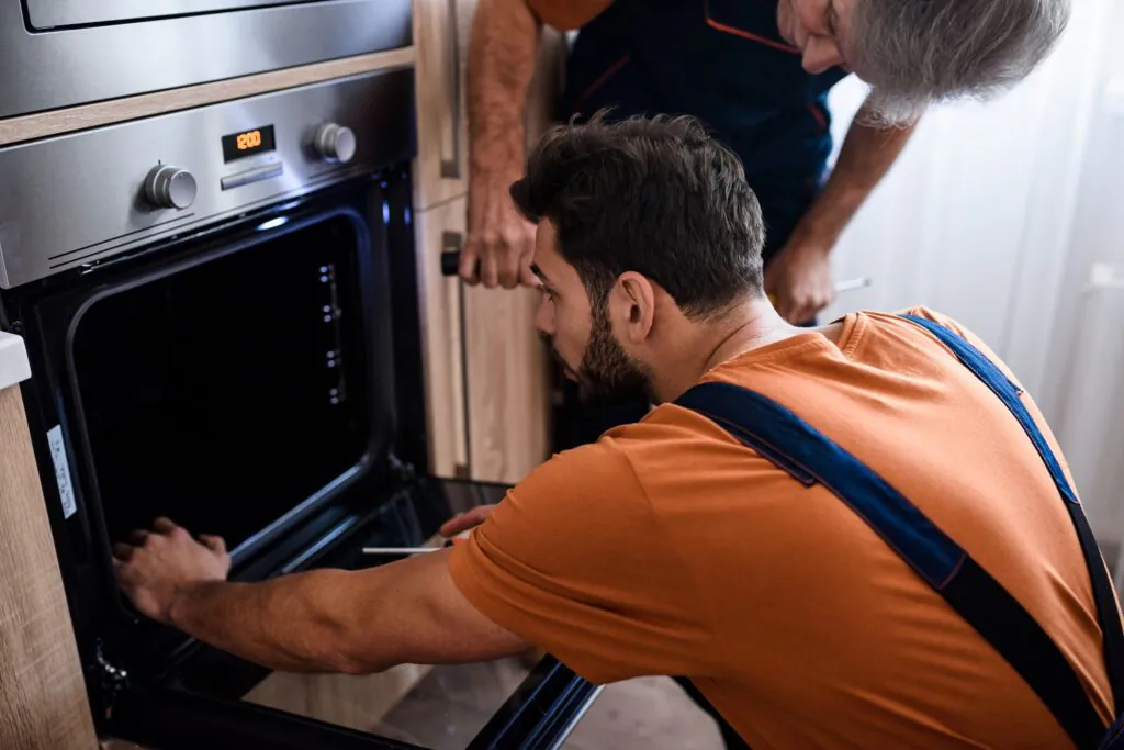 A male technician inspects an open oven, assisted by another person, performing field service management in a modern kitchen. The oven display reads "12:00".