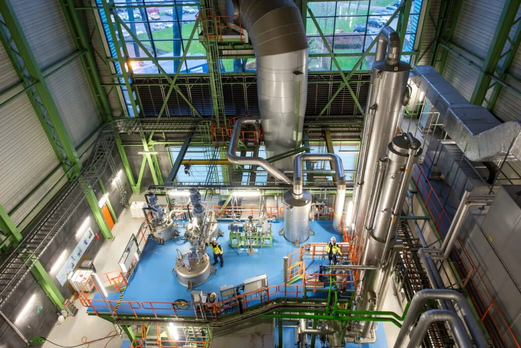 Two workers perform **field service management** on complex industrial processing equipment in a modern, high-ceiling plant. Visible text: "Bauman Group".