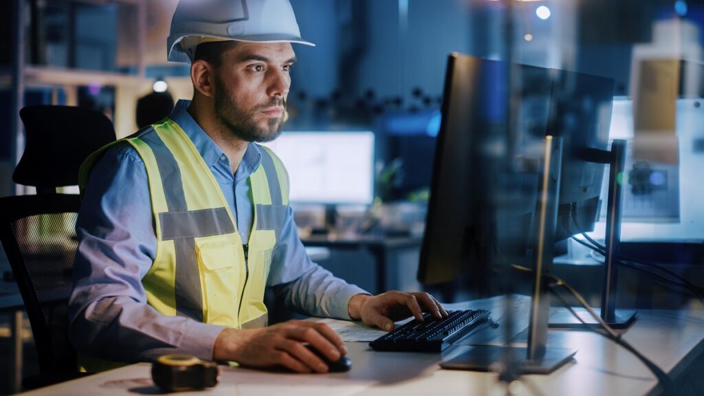 Een technicus met helm en veiligheidsvest werkt achter een computer aan optimalisatie van asset management binnen een kantooromgeving met meerdere schermen in de avond.