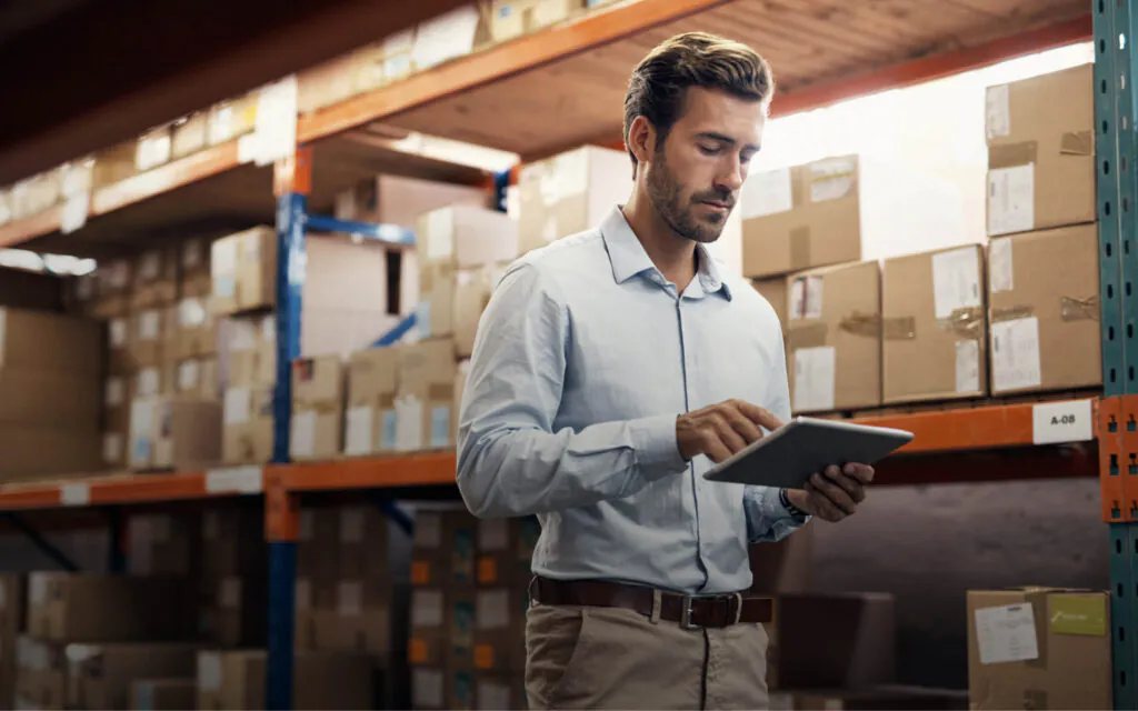 Een man in een lichtblauw overhemd bedient een tablet in een magazijn vol kartonnen dozen, wat het proces van asset management ondersteunt. Tekst op het label: A-08.