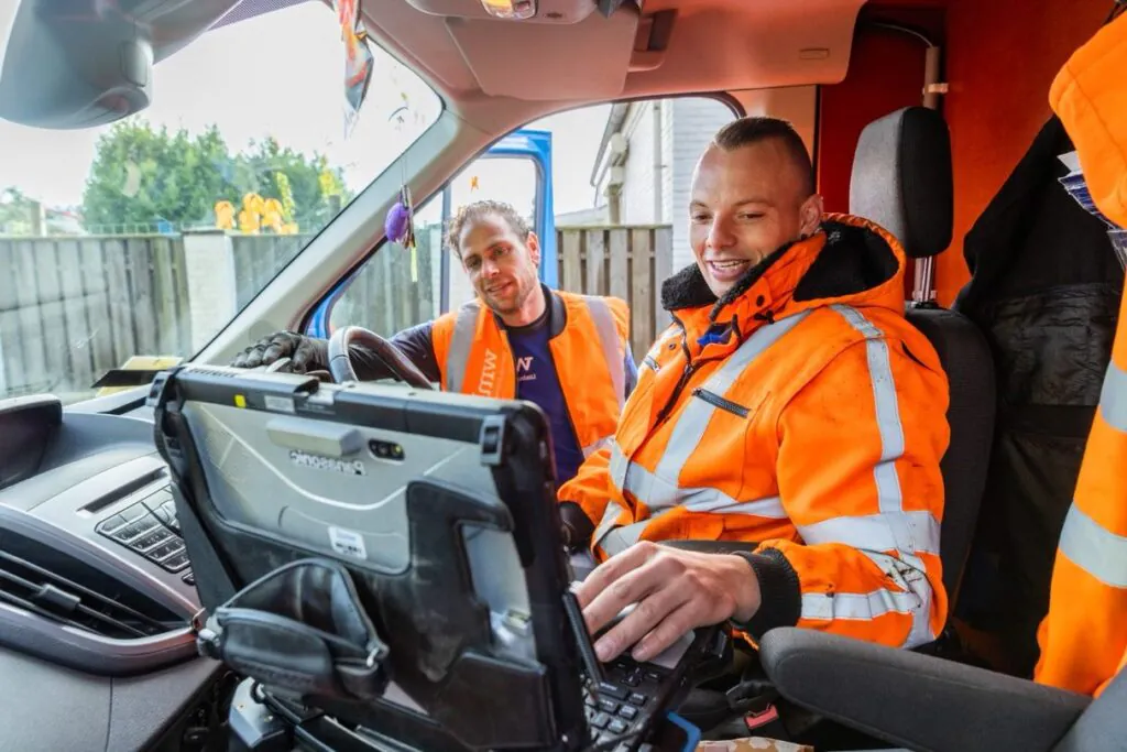 Twee monteurs in oranje reflecterende werkkleding bedienen een Panasonic-laptop in de cabine van een bedrijfswagen voor optimale field service management, terwijl ze buiten een woonwijk observeren.
