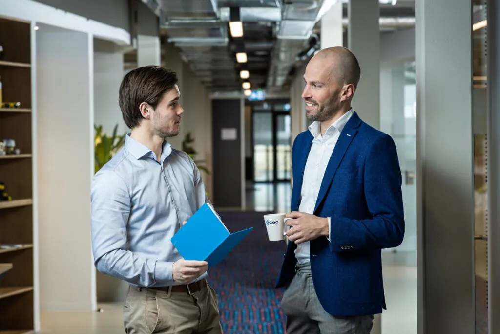 Twee collega's bespreken projecten en SAP FSM-oplossingen terwijl ze praten in een moderne kantoorhal. De man rechts houdt een mok vast met de tekst: "ideo".