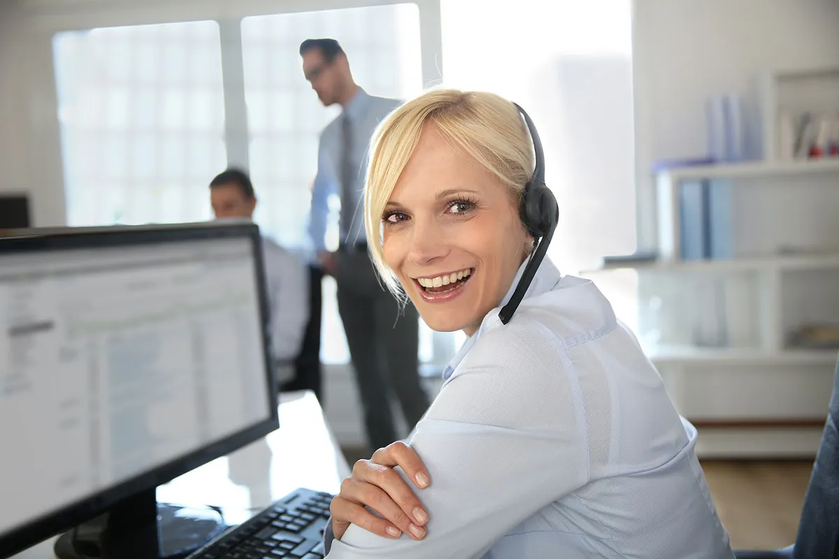 Een glimlachende vrouwelijke medewerker met een headset kijkt achterom terwijl ze SAP FSM gebruikt aan haar bureau in een kantooromgeving met collega's op de achtergrond.