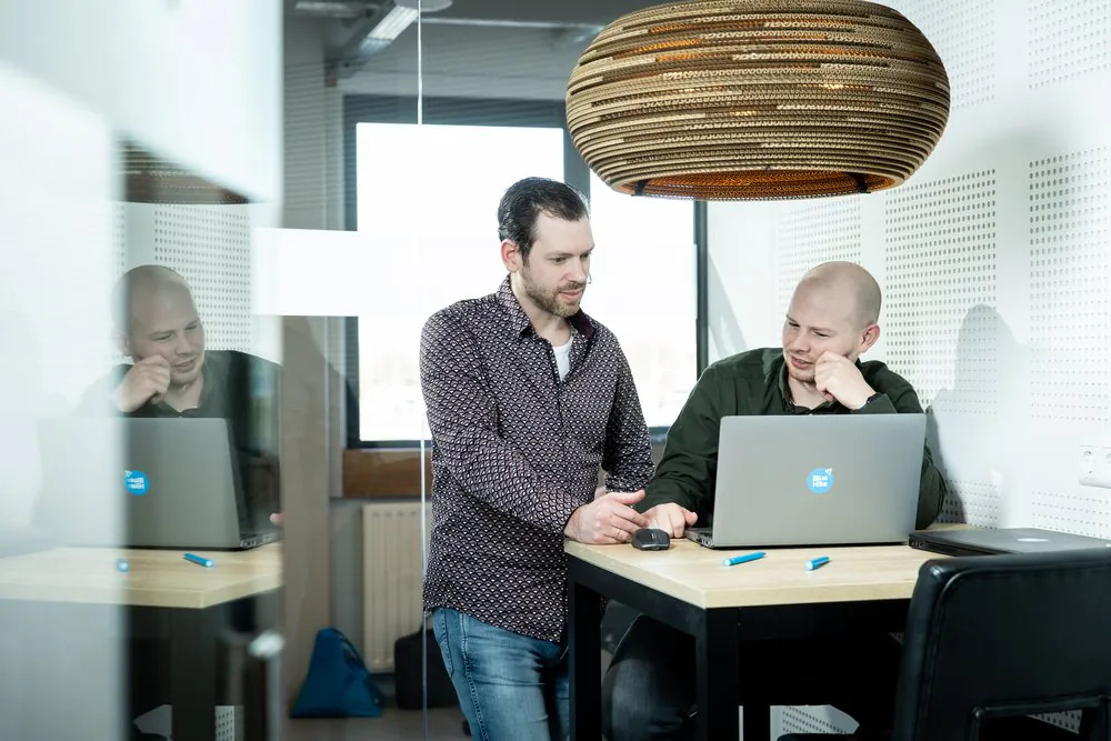 Twee mannen werken aan een laptop en bespreken sap fsm in een modern kantoor met een opvallende hanglamp en een lichte houten tafel. Tekst op laptop: Blue Hike.