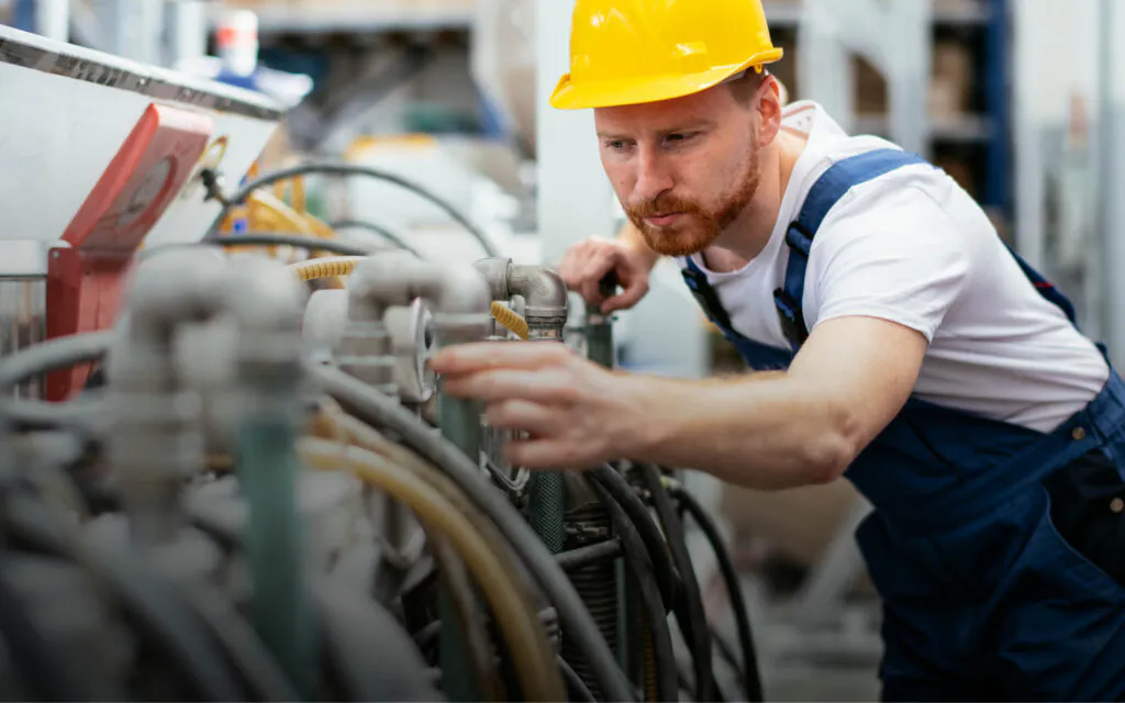 Een monteur met een gele helm inspecteert geconcentreerd industriële machines voor effectief field service management in een fabriekshal, waarbij hij nauwkeurig verschillende leidingen en onderdelen van de installatie controleert.