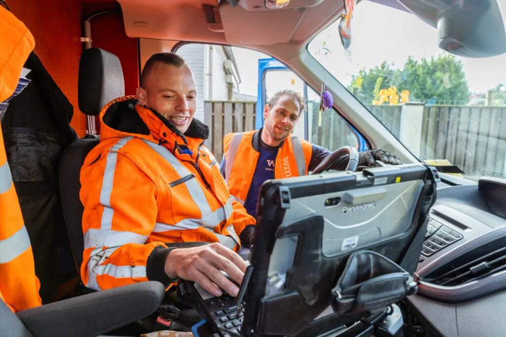 Twee monteurs in oranje werkkleding gebruiken een Panasonic-tablet voor field service management terwijl ze in een bestelwagen zitten en samen naar het scherm kijken.