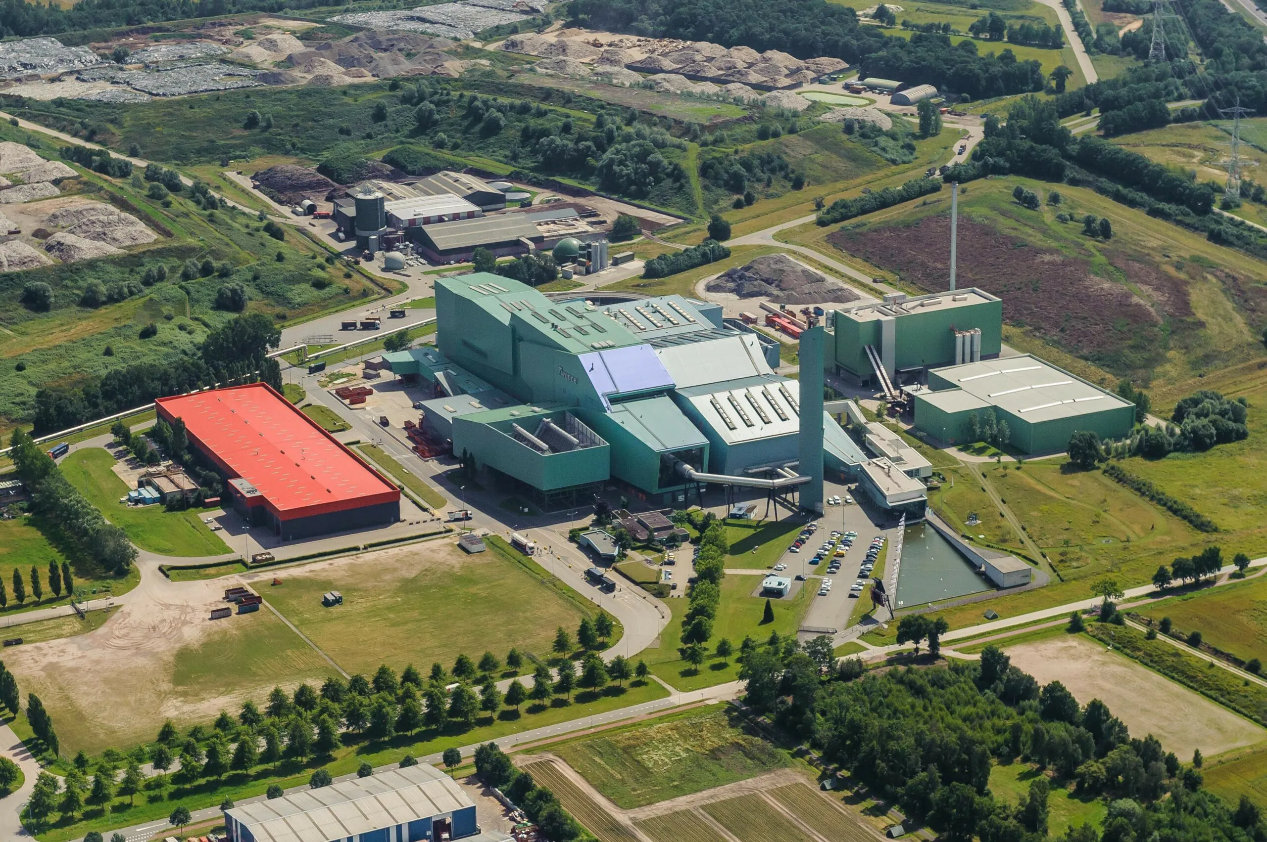 Een industrieel complex met groene en rode daken staat in een landelijke omgeving, waarbij de infrastructuur efficiënt wordt onderhouden met sap fsm voor optimaal asset management. Tekst: Twence.