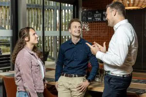 Drie collega’s praten glimlachend met elkaar in een kantooromgeving terwijl ze ideeën uitwisselen over SAP FSM. Tekst op de achtergrond: EL COFFEE TIME.