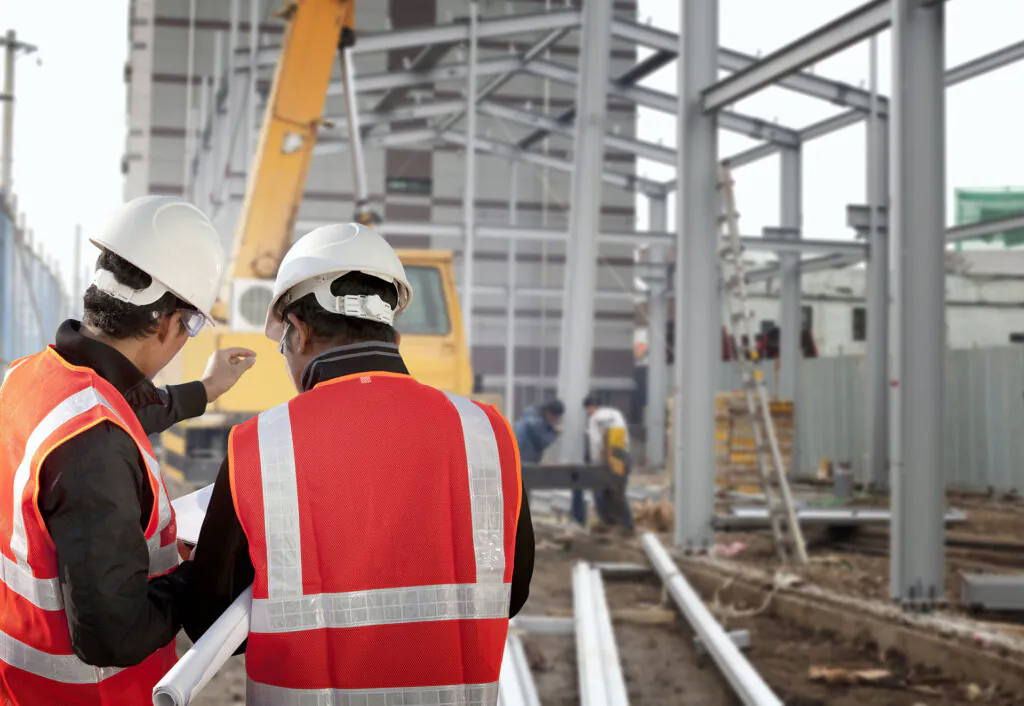 Twee bouwvakkers in veiligheidsvesten en helmen bespreken bouwplannen op een bouwplaats, ter ondersteuning van efficiënt field service management bij het realiseren van de staalconstructie op de achtergrond.