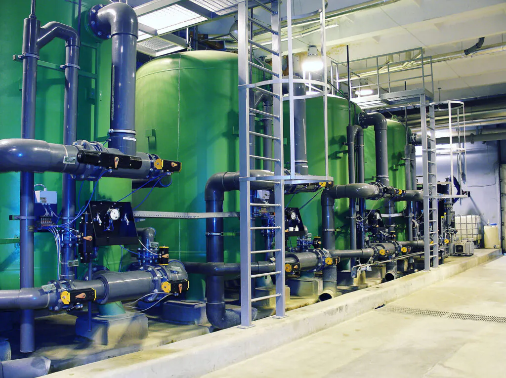 Grote groene tanks en grijze leidingen voor asset management staan in een industriële faciliteit, verbonden door een complex buizensysteem en toegankelijk via een verticale metalen ladderconstructie in de ruimte.