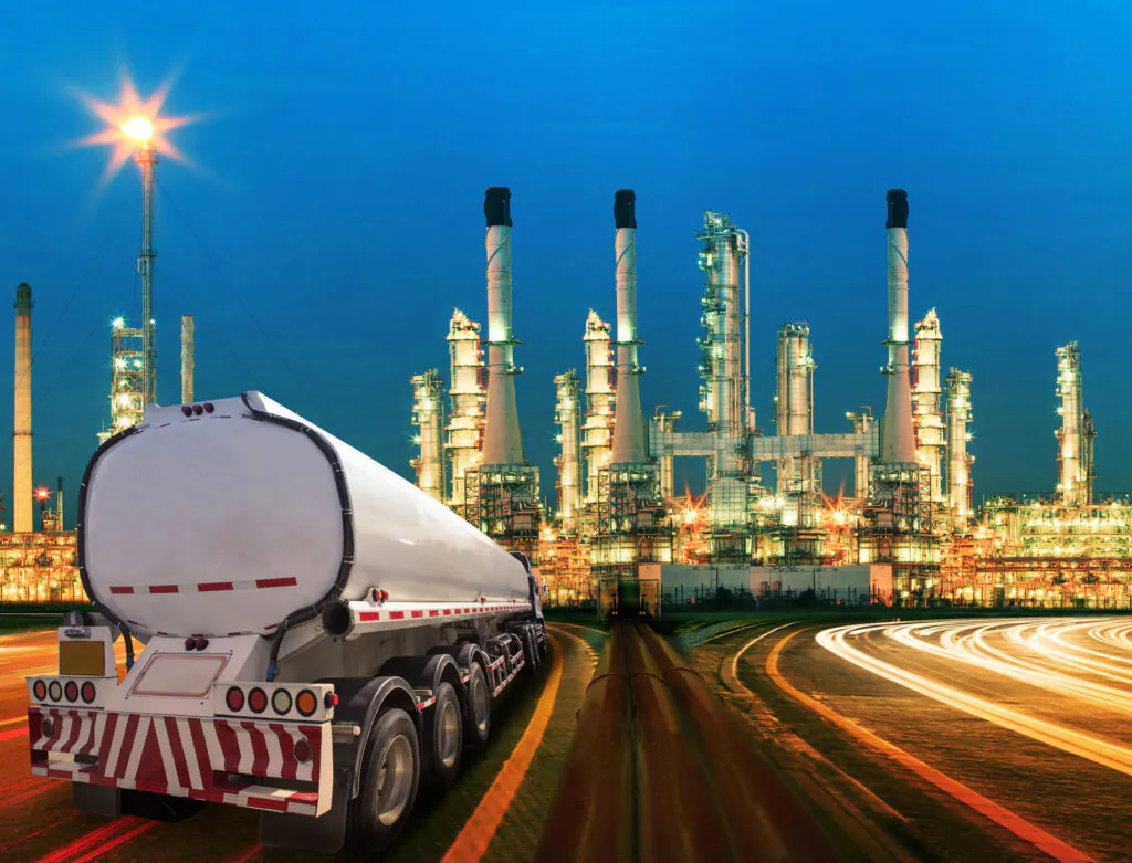 Een tankwagen rijdt op een weg langs een verlichte olieraffinaderij bij schemering, waarbij effectieve asset management cruciaal is voor de operationele processen en efficiëntie in deze industriële omgeving.