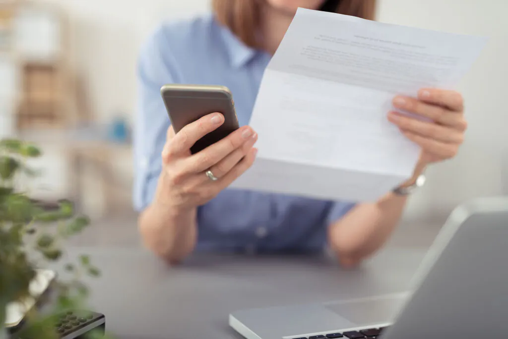 Een vrouw houdt een smartphone en een document vast, wat helpt bij efficiënt asset management op een kantoorwerkplek met een laptop op de voorgrond. Tekst op het document: "as soon as possible. Please let us know if you have any questions or concerns. We look forward to hearing from you. Kind regards,