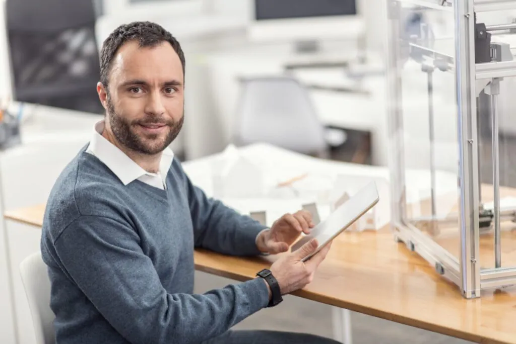 Een man met baard bedient een tablet in een kantooromgeving met een 3D-printer, wat bijdraagt aan efficiënt asset management en de optimalisatie van moderne werkprocessen.