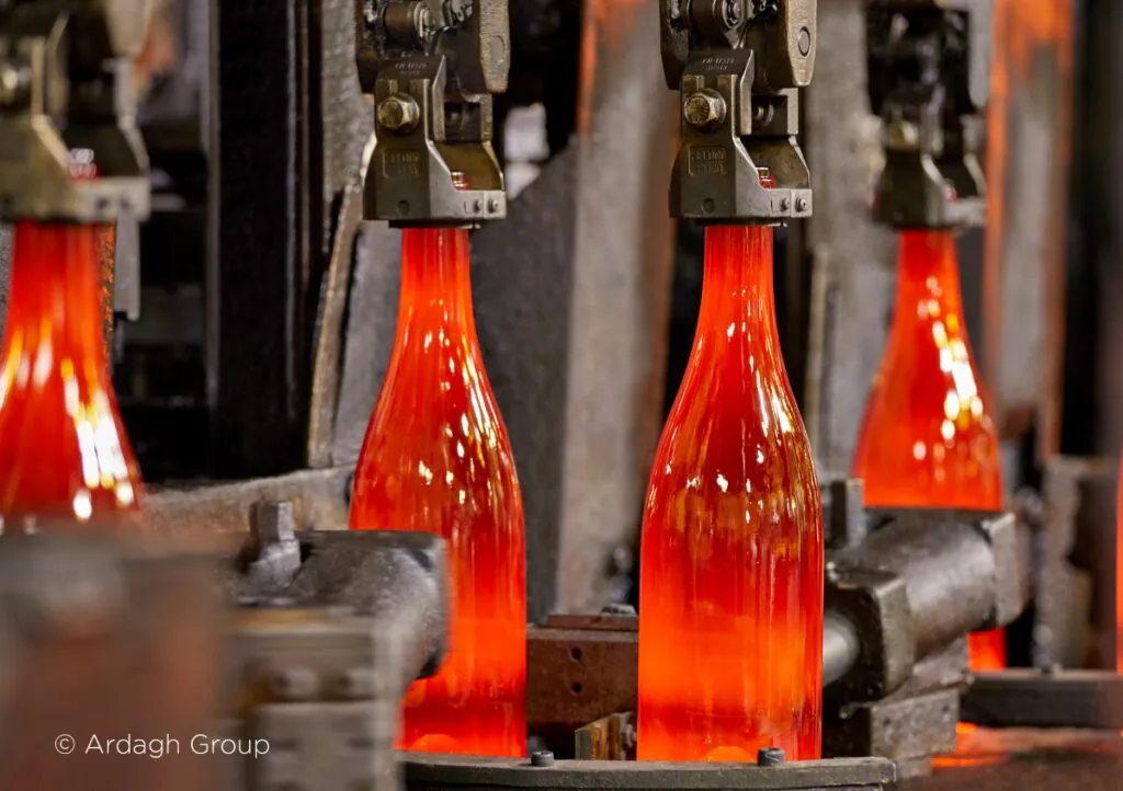 Gloeiende glazen flessen worden door mechanische grijpers langs een productielijn getransporteerd voor optimaal asset management in een industriële glasfabriek. Tekst op de afbeelding: © Ardagh Group