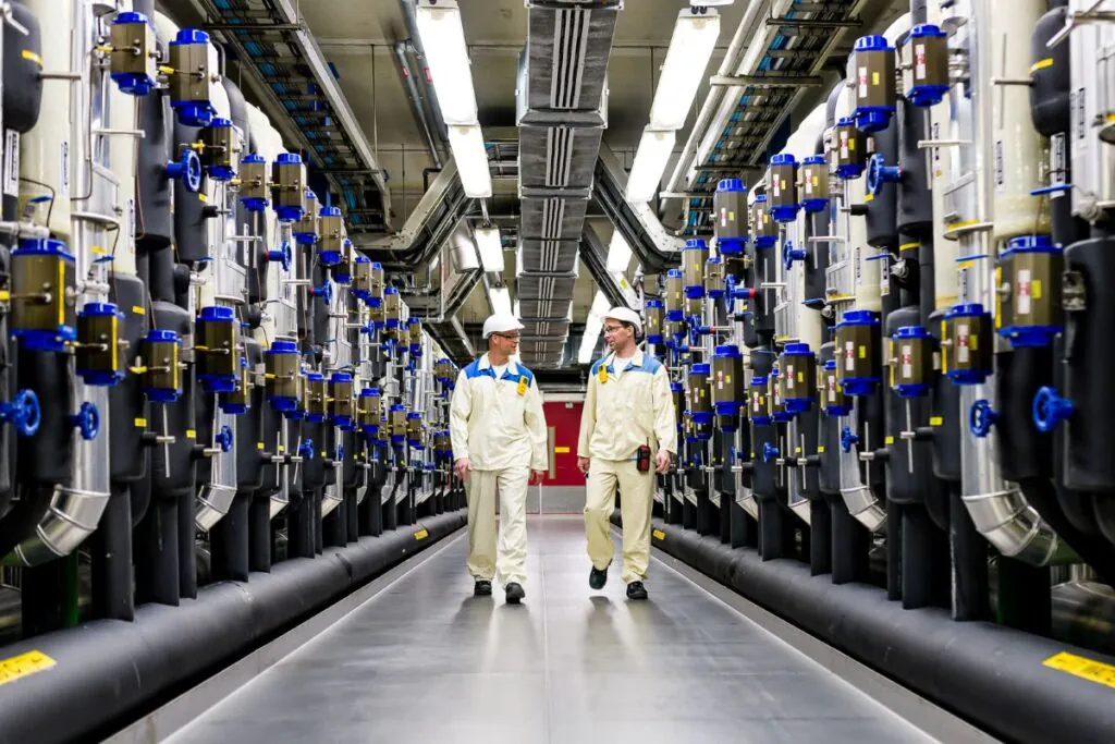 Twee technici in werkkleding lopen door een industriële gang tussen rijen complexe leidingen en machines, ter ondersteuning van efficiënt field service management in een technische omgeving.